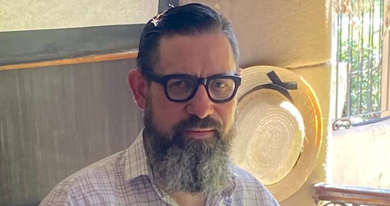 Lew MacDonald is wearing dark rimmed glasses and a white collared shirt. He has a full bear and dark hair. 