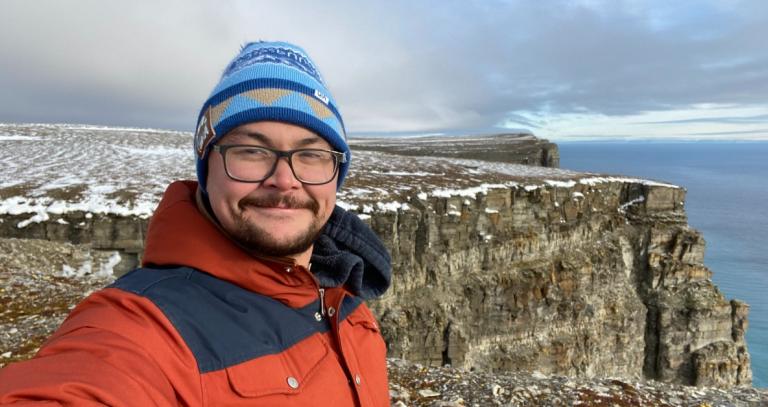 Josh Komangapik, smiling and standing on a stone cliff wearing an orange and blue coat and blue hat. The ocean is visible beyond the cliff.