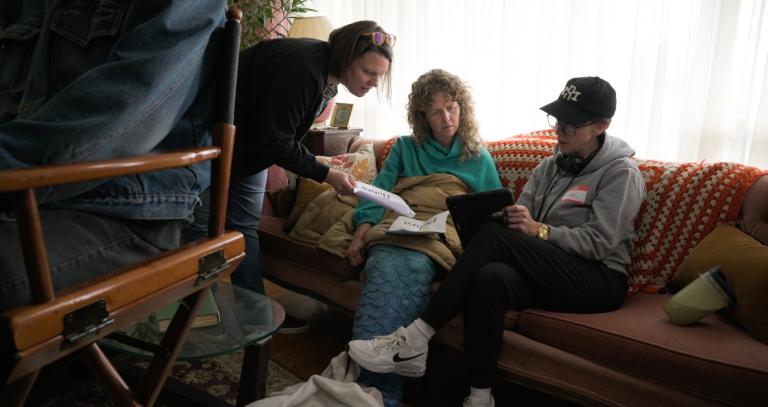 Kim Manky handing papers to a woman sitting on sofa wearing a mermaid costume.