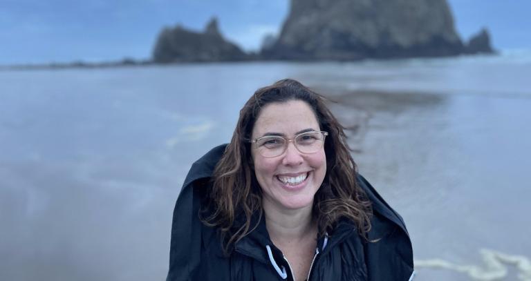 Kyle Epstein smiling and standing on the shore of a body of water with a large rock in the background offshore.