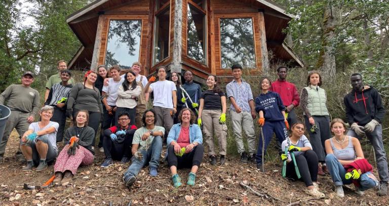 Group of smiling students from Pearson College UWC standing in front of a cabin in the forest.