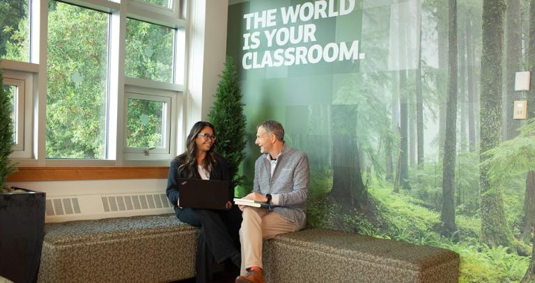 A woman and man sitting and talking with "The World is your classroom" on the wall behind them.