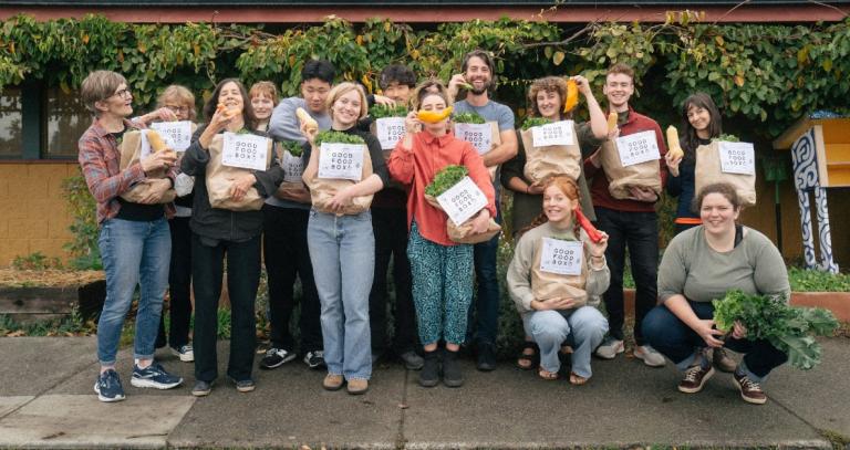 Group of people holding Fernwood Neighbourhood House Good Food bags. They are holding up different fruits and vegetables and smiling.