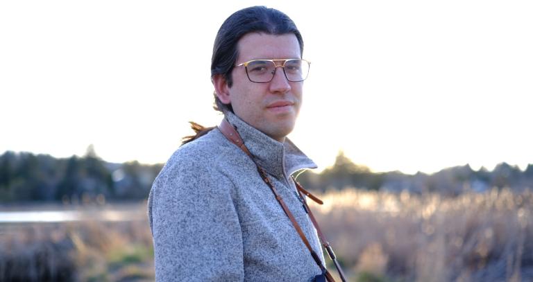 Jeff Nicholls is looking at the camera with trees, water, and low brush vegetation in the background.