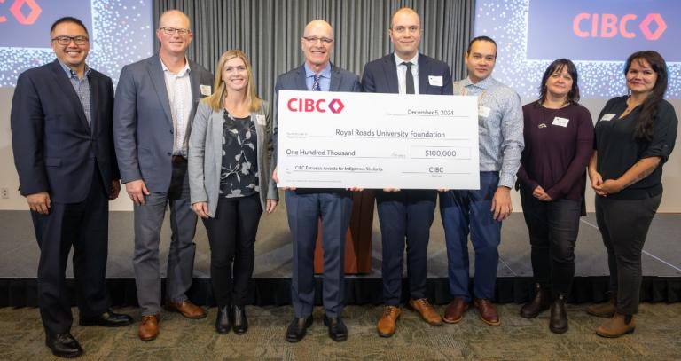 Nelson Chan, Matt Gillberg, Jacky Weber, Philip Steenkamp, Spencer Walker, Russell Johnston, Cherylanne James, Shelby Blackjack stand in a line. Philip and Spencer hold a large cheque for $100,000 from CIBC to Royal Roads University.