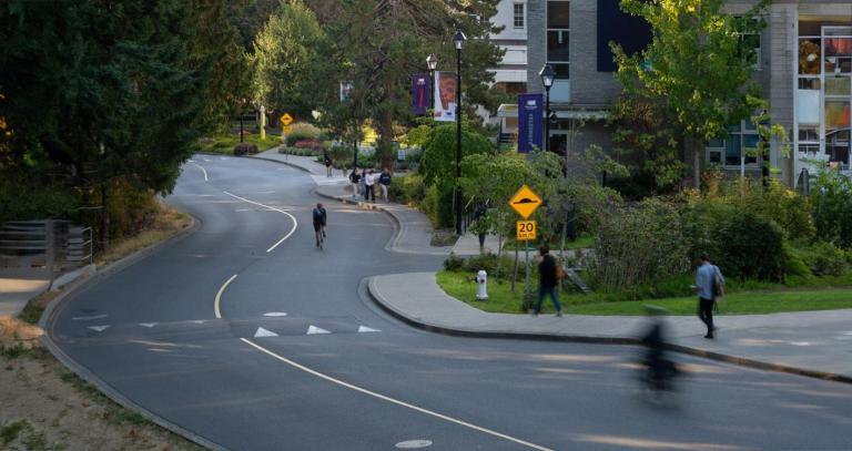 Bikers and walkers wind down a paved road alongside green foliage at Royal Roads University