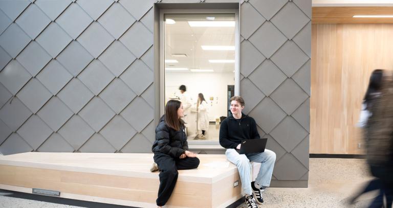Two students sit on a wooden bench in front of a lab inside an RRU building
