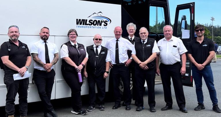 Group of smiling employees in front of a passenger bus with Wilson's logo on the side of it.