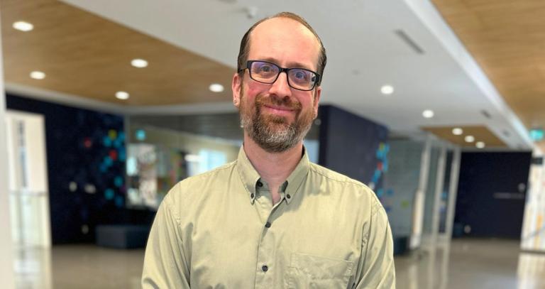A man in glasses, poses for a headshot in a hallway.