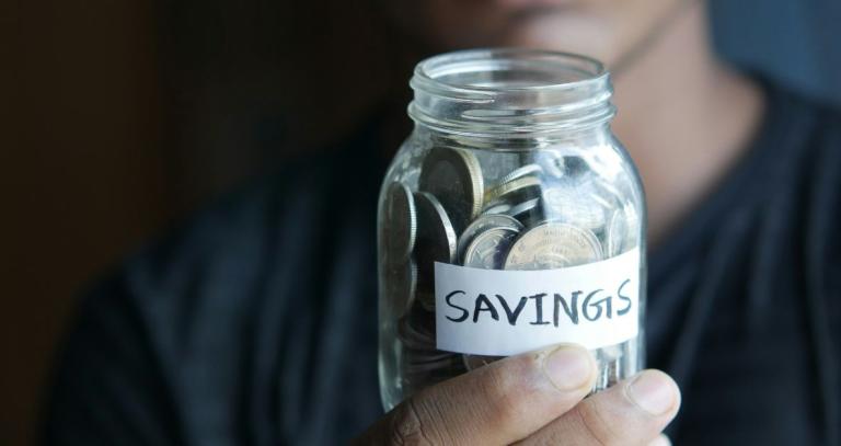 A hand holding a jar labeled "savings" with coins inside