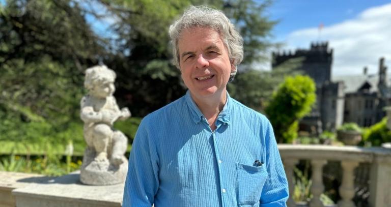 A man in a blue collared shirt smiles. A cherub statue is visible in the background.