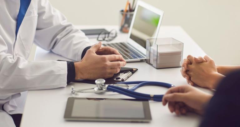 A doctor sitting at a desk with their hands on the table in front of them, with a computer open and a patient across from them. 