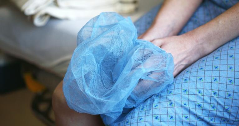Person's lap shown sitting on hospital bed holding a hospital cap in their hands.