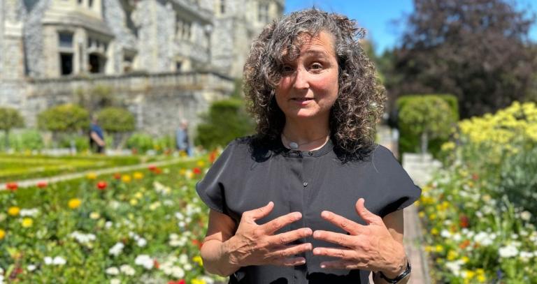 A woman stands in front of a garden and castle, her hands gesturing as she speaks