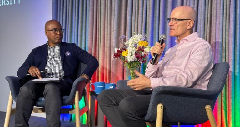 Two men on stage in conversation. Rainbow lights can be seen lighting the background.