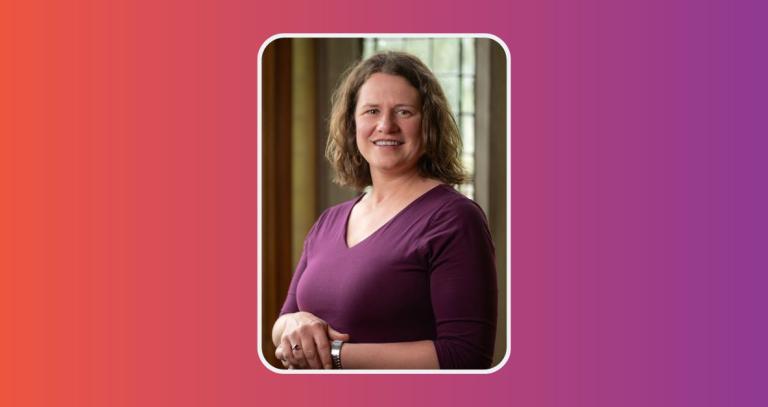 Eva Malisius smiling wearing a purple sweater with a glass window and wood panel walls behind her. 