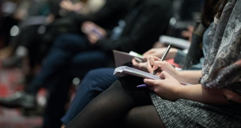 Woman holding a notebook in her lap and taking notes, her face is not seen in the image.