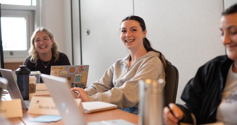 Three young women sitting at a table with laptops open and smiling.