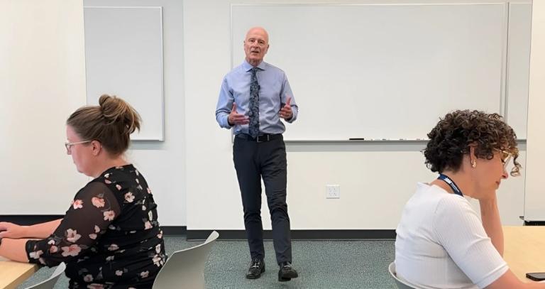 A man stands in a classroom with students reading at tables on either side.