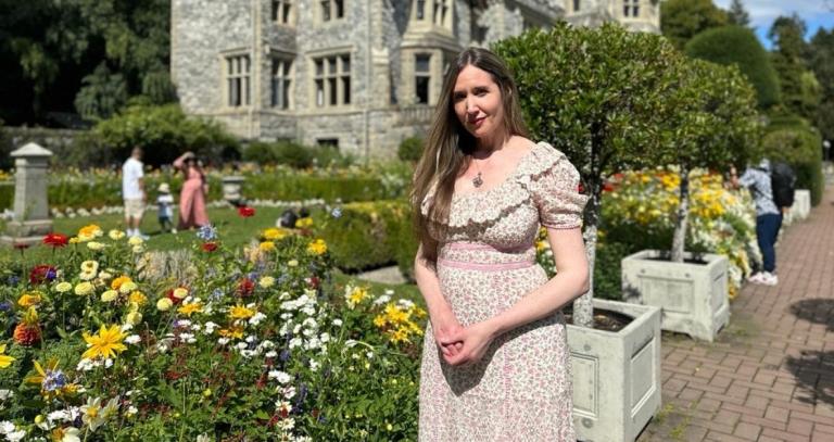 A woman poses in a colourful flower garden, with a stone castle in behind her.
