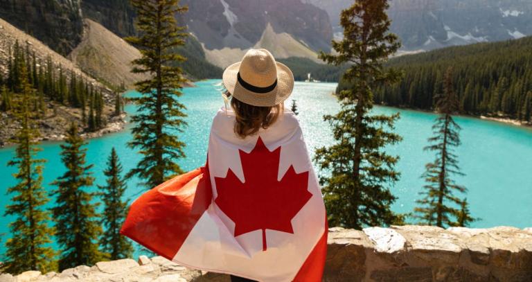 A woman stands wrapped in a Canadian flag in front of a blue lake and mountains