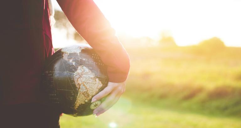 Person holding a globe by their side with trees and sunshine in the background.