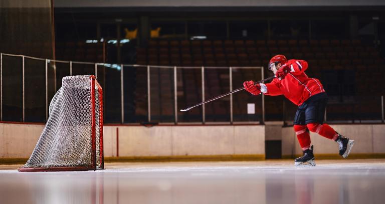 A man in hockey attire on an ice rink hitting a puck into the net. 