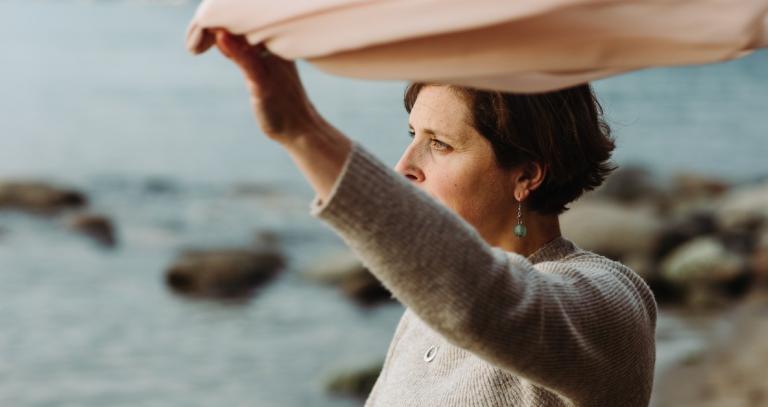 Cheryl Heykoop standing on a beach holding a pink scarf high in the wind.