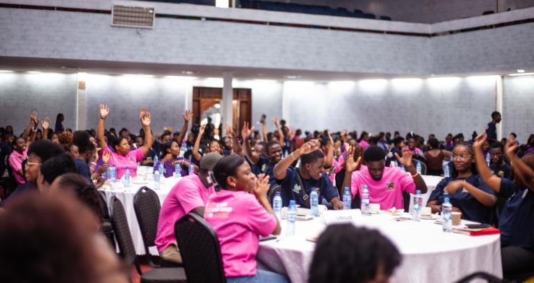 groups of people seated at round tables wearing hot pink t-shirts, their hands are raised.