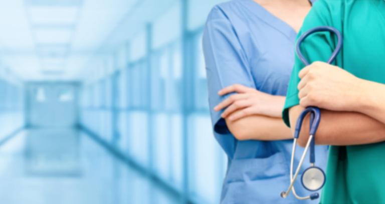 Two doctors shown in scrubs, one is holding a stethoscope, standing in a long glass hallway.