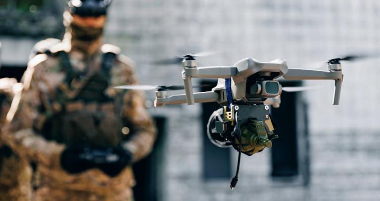 A soldier in combat fatigues using a handheld console to fly a drone, featured in the foreground