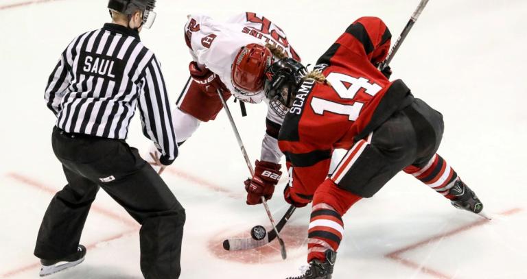 Two women hockey players face off. A ref drops a puck.