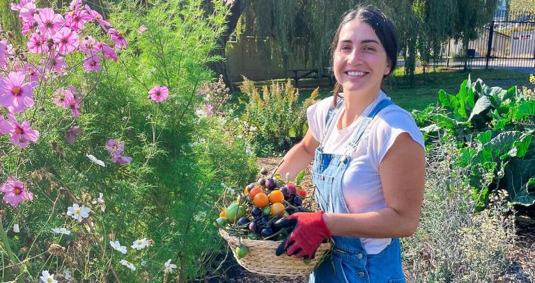 Emily Mulroney holding a basket of vegetables standing by tall flowers in The Farm at RRU