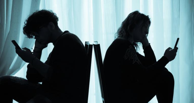 Two teenagers in a dark room, back to back, staring at their smartphones