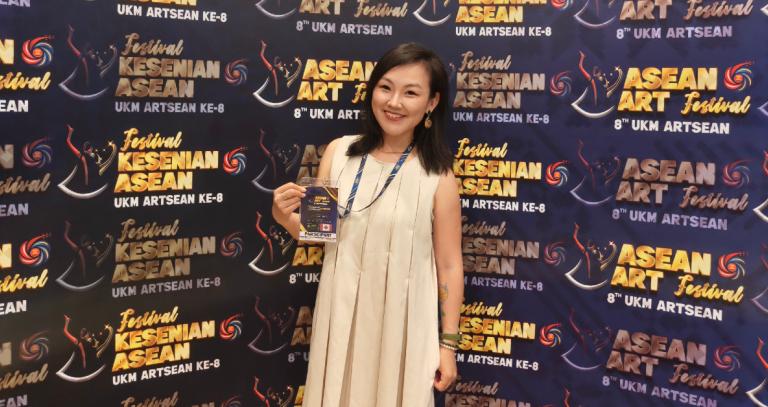 Shuwen Tian holding up her delegate badge and standing in front of ASEAN ART Festival 8th UKM Artsean backdrop