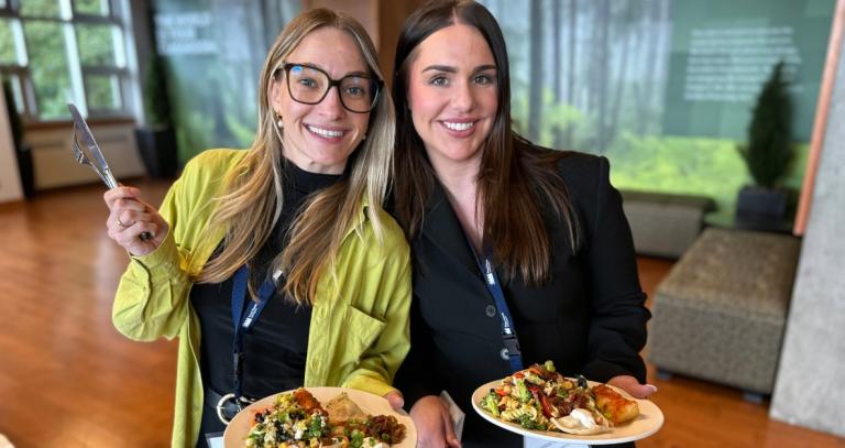 Two students smile at the camera while holding plates filled with perogies, cabbage rolls and salad.