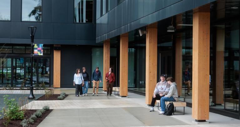 Groups of students can be seen sitting and walking on a university campus.