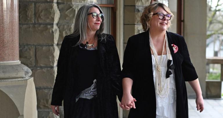 Two woman look over their shoulder while holding hands.