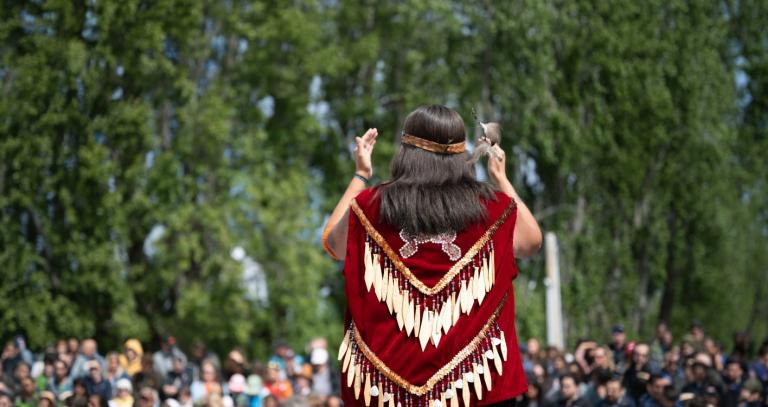 An Indigenous performer raises her hands during a performance as seen from behind.