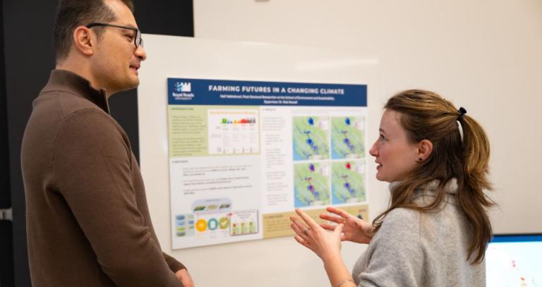 A woman gestures while speaking to a man, a poster board behind them.