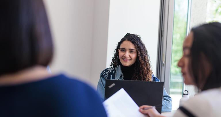 RRU students sit around a table by a large window with their laptops and engage in conversation