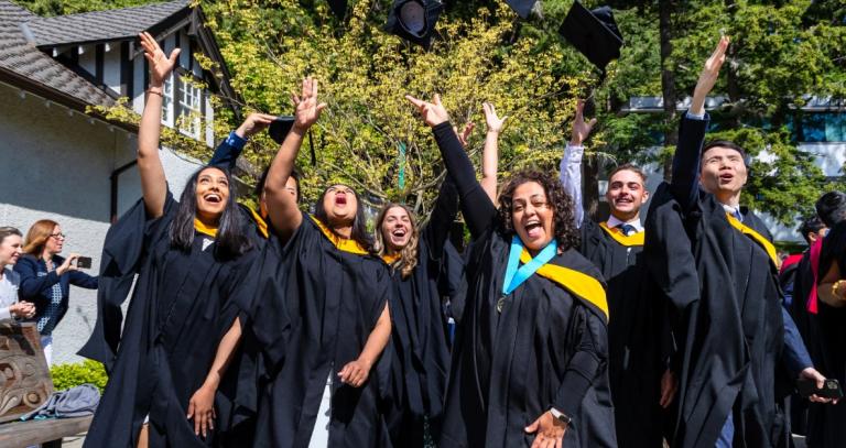 A group of graduates in convocation robes with yellow sashes throw their grad caps in the air and smile