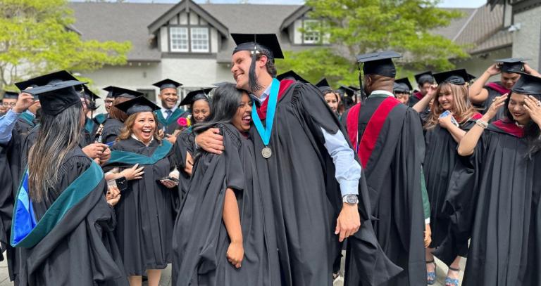 Two graduates hug after convocation with a bunch of others wearing caps and gowns gather behind them.