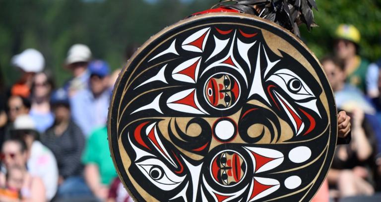 A Lekwungen dancer holds a round drum with Coast Salish art on it