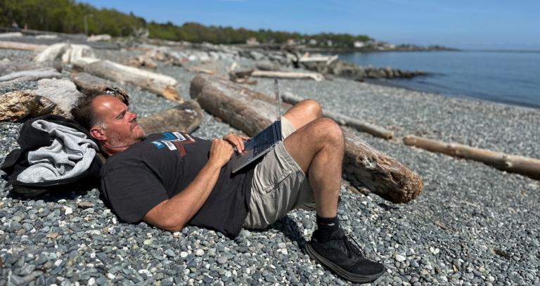 A man lays on a rocky beach, surrounded by driftwood. He's laying down, working on his laptop.