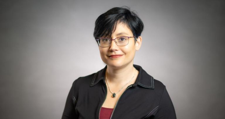 A studio head shot of a woman with a gentle smile in front of a grey screen. She has short, cropped hair.