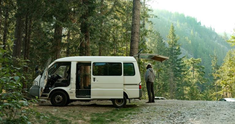 A camper stands beside their van in a forested area