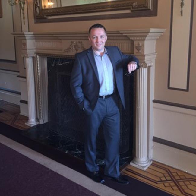 Royal Roads University alumni stands in the castle next to a fireplace with his arm on the mantle