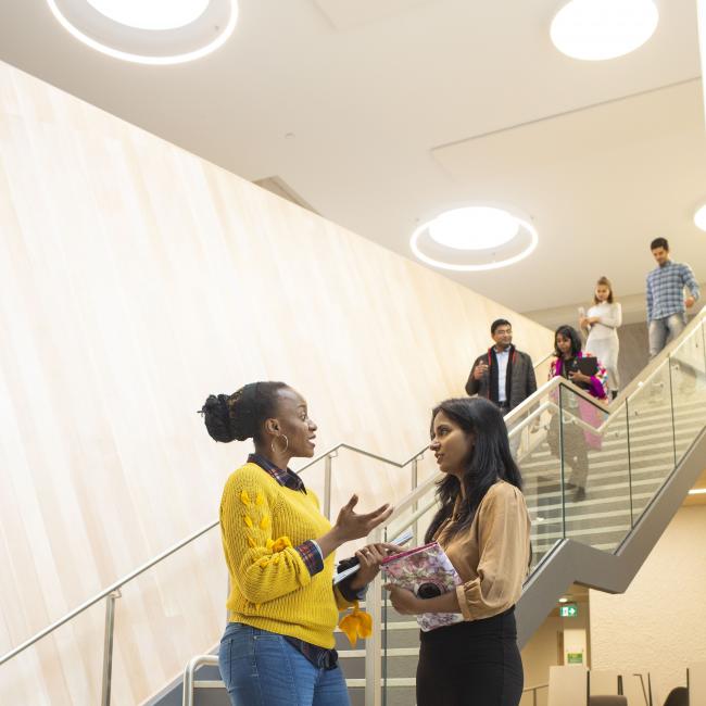 Two students chat at the bottom of a brightly lit staircase that several others are descending.