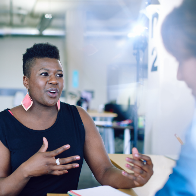 A young black woman gestures to a classmate as though describing something she is passionate about. A spark of light behind her speaks to a bright future ahead.